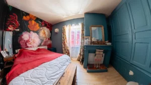 Chambre décorée avec papier peint floral, lit au couvre-lit rouge, murs bleus et miroir au-dessus d'une cheminée